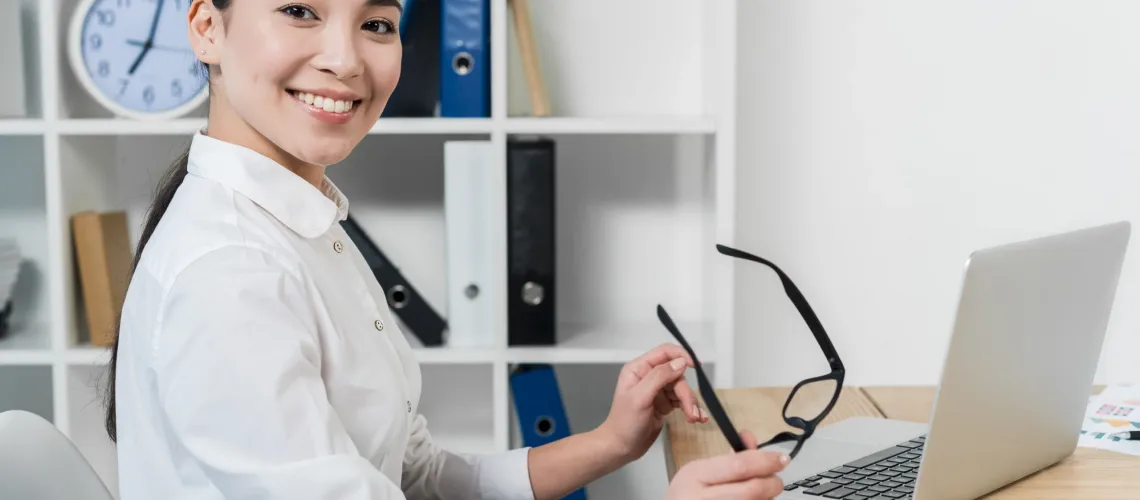 portrait-smiling-young-businesswoman-holding-eyeglasses-hand-with-laptop-desk