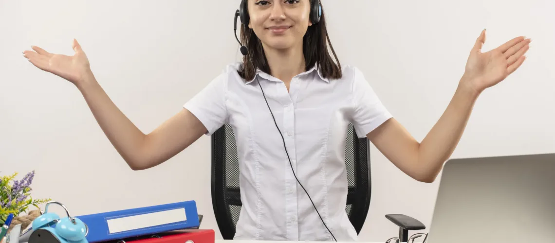 young girl in white shirt and headphones with a microphone looking at camera with smile on face spresding hands to the sides sitting at the table with folders and laptop over white background